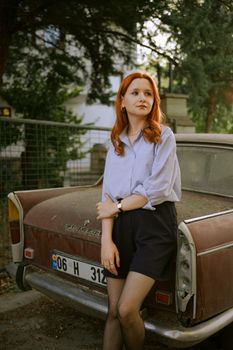 A stylish woman with red hair poses by a vintage Peugeot car in an urban setting, exuding elegance.