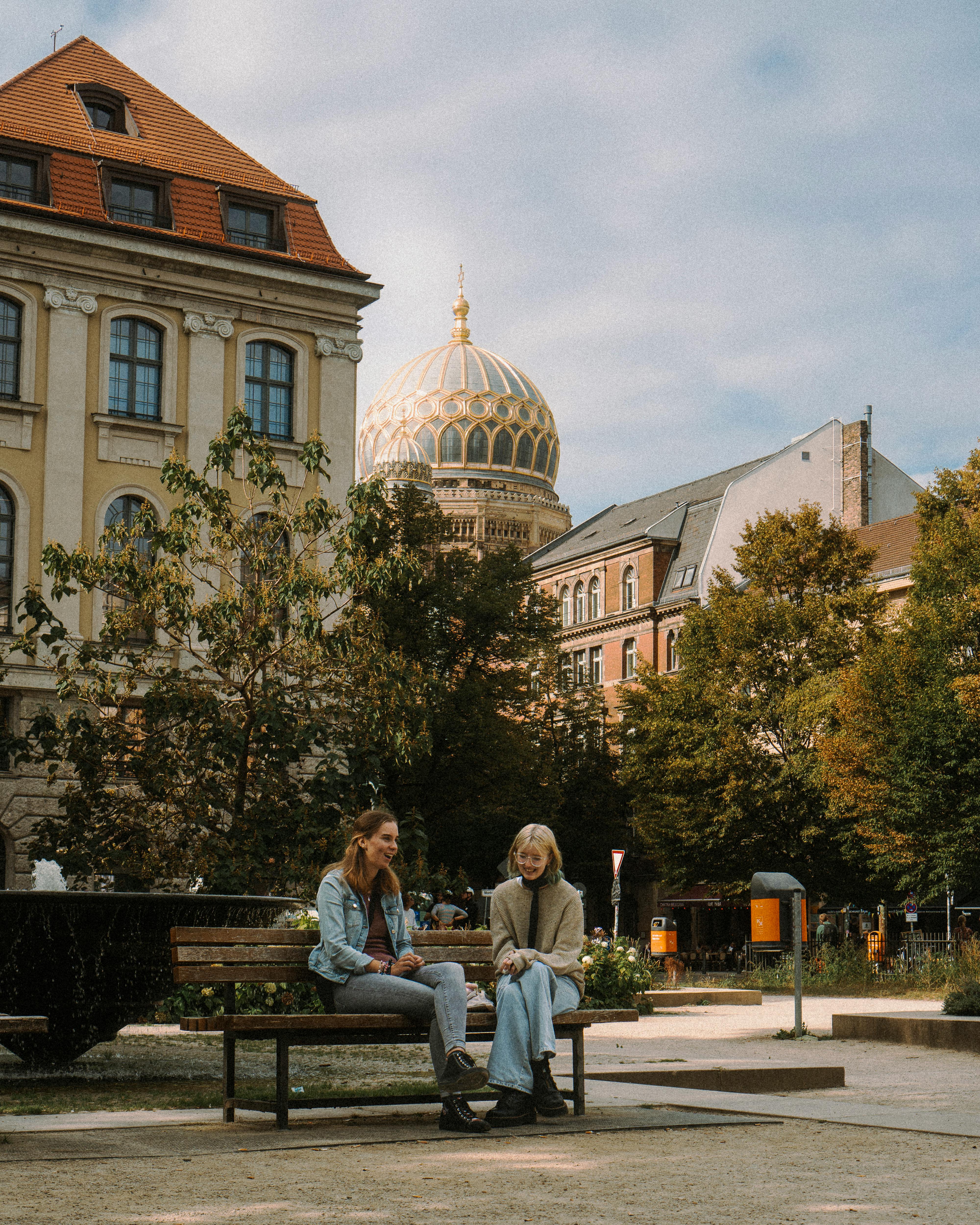 Two women enjoying a sunny day on a bench in a Berlin park with historic architecture nearby.