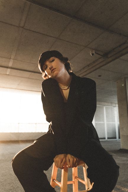 A woman in a beanie and black jacket poses on a stool in a parking garage, evoking a moody atmosphere.