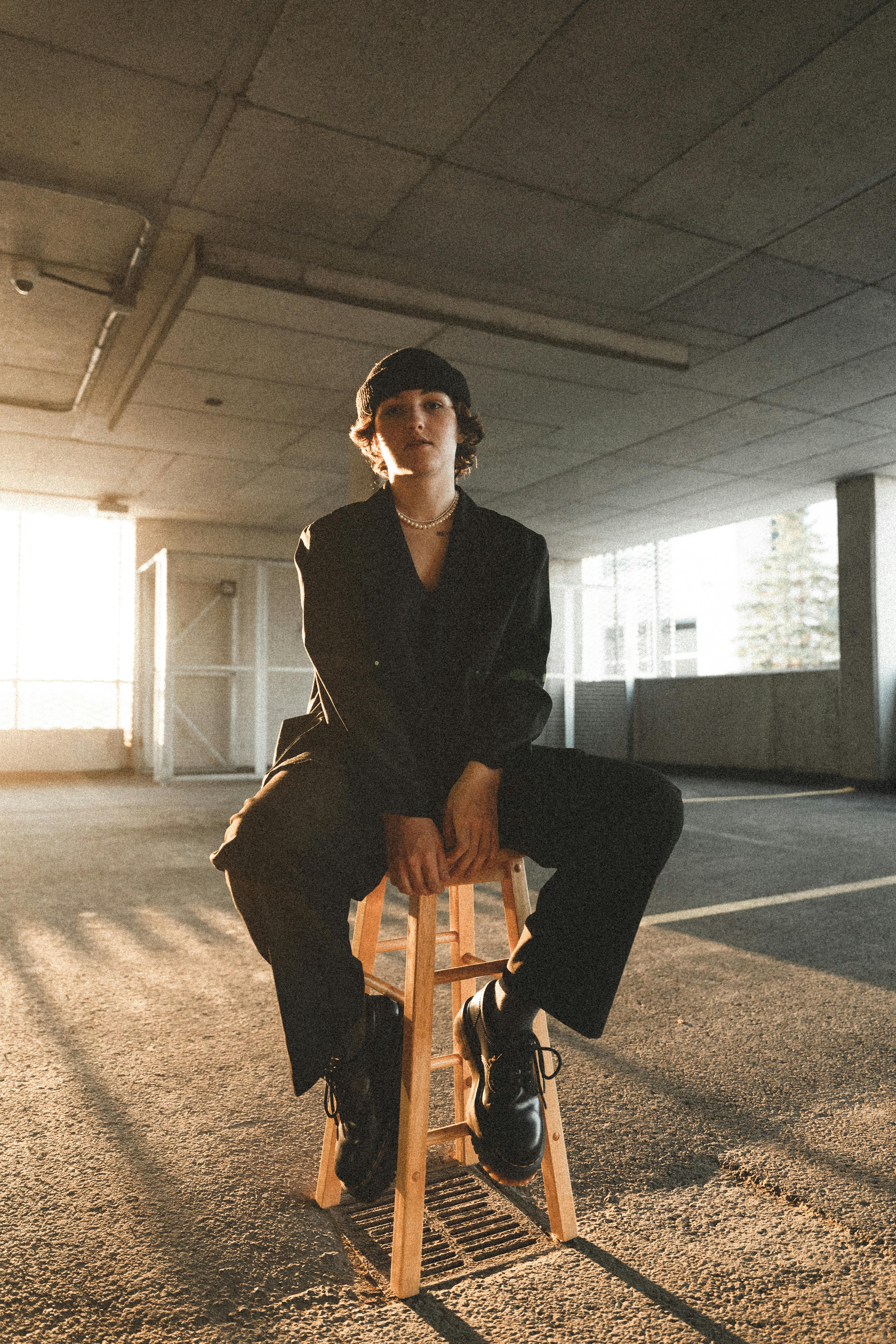 A stylish woman in black attire sits on a stool in an industrial parking garage, capturing urban fashion vibes.