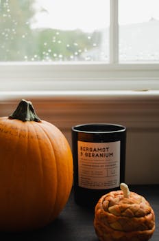 An autumn-themed interior featuring a pumpkin and a scented candle by a rainy window.