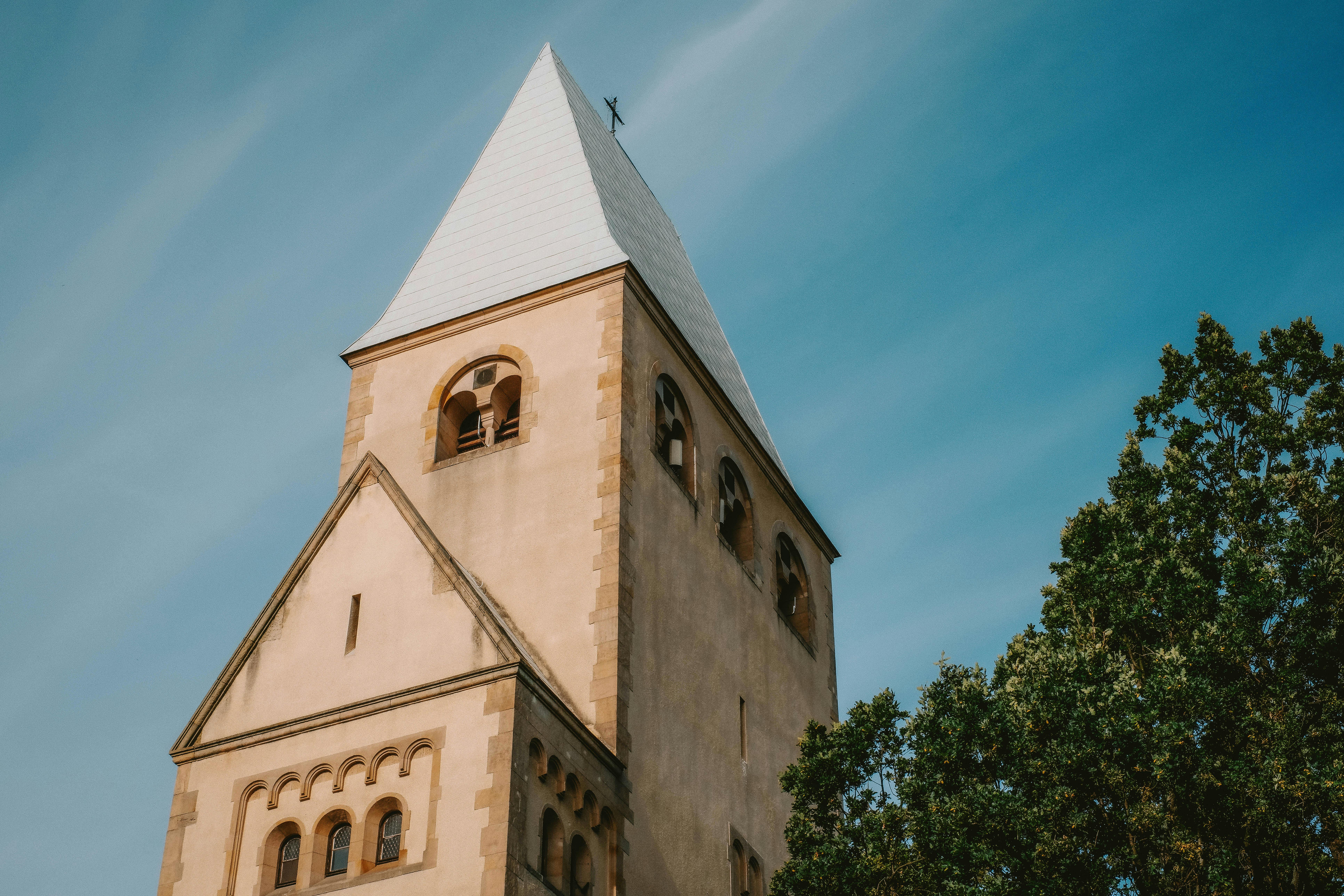 Low-angle view of a historic church tower with a tree in the foreground and a clear blue sky.