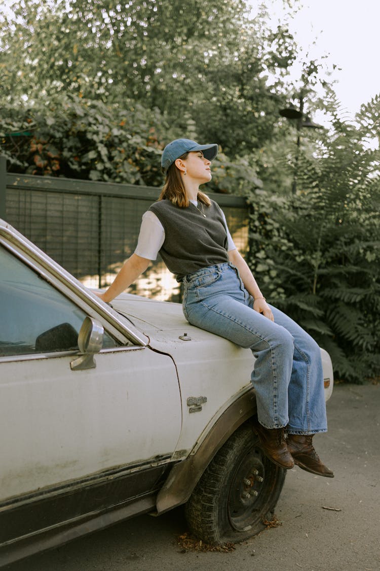 Woman In Cap Sitting On Car And Posing