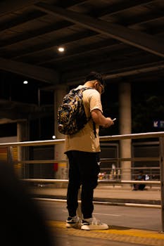 Man with backpack using smartphone on city street at night, creating an urban scene.
