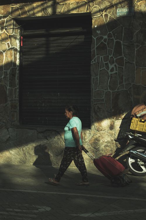 Free A woman walks in the city pulling a suitcase, shadows casting against a stone wall. Stock Photo