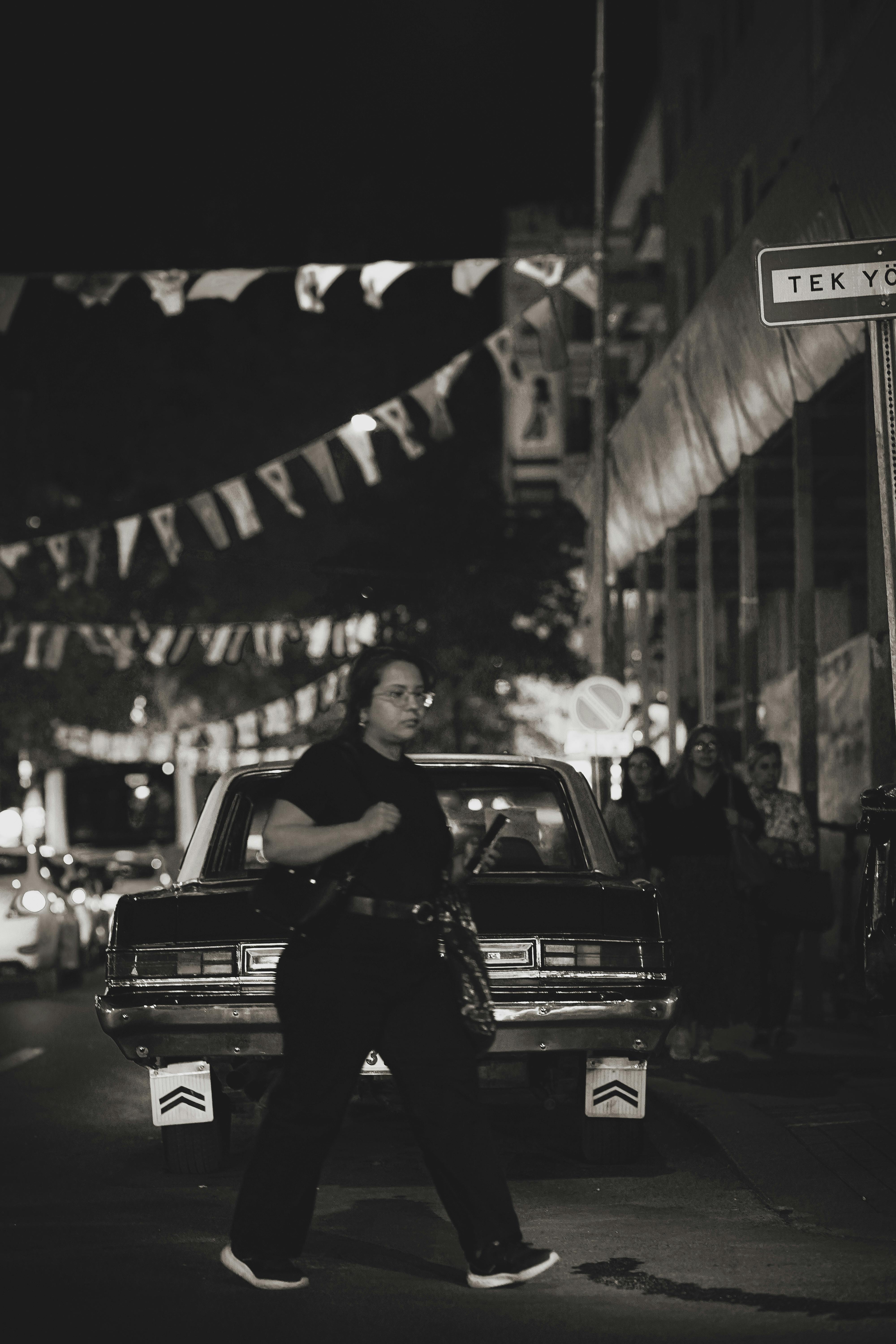 Black and white photo of a woman crossing city streets in Ankara at night with a vintage car.