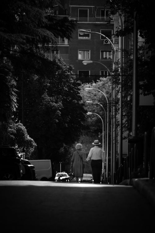Free Elderly couple walking hand in hand down a shadowed urban street in black and white. Stock Photo
