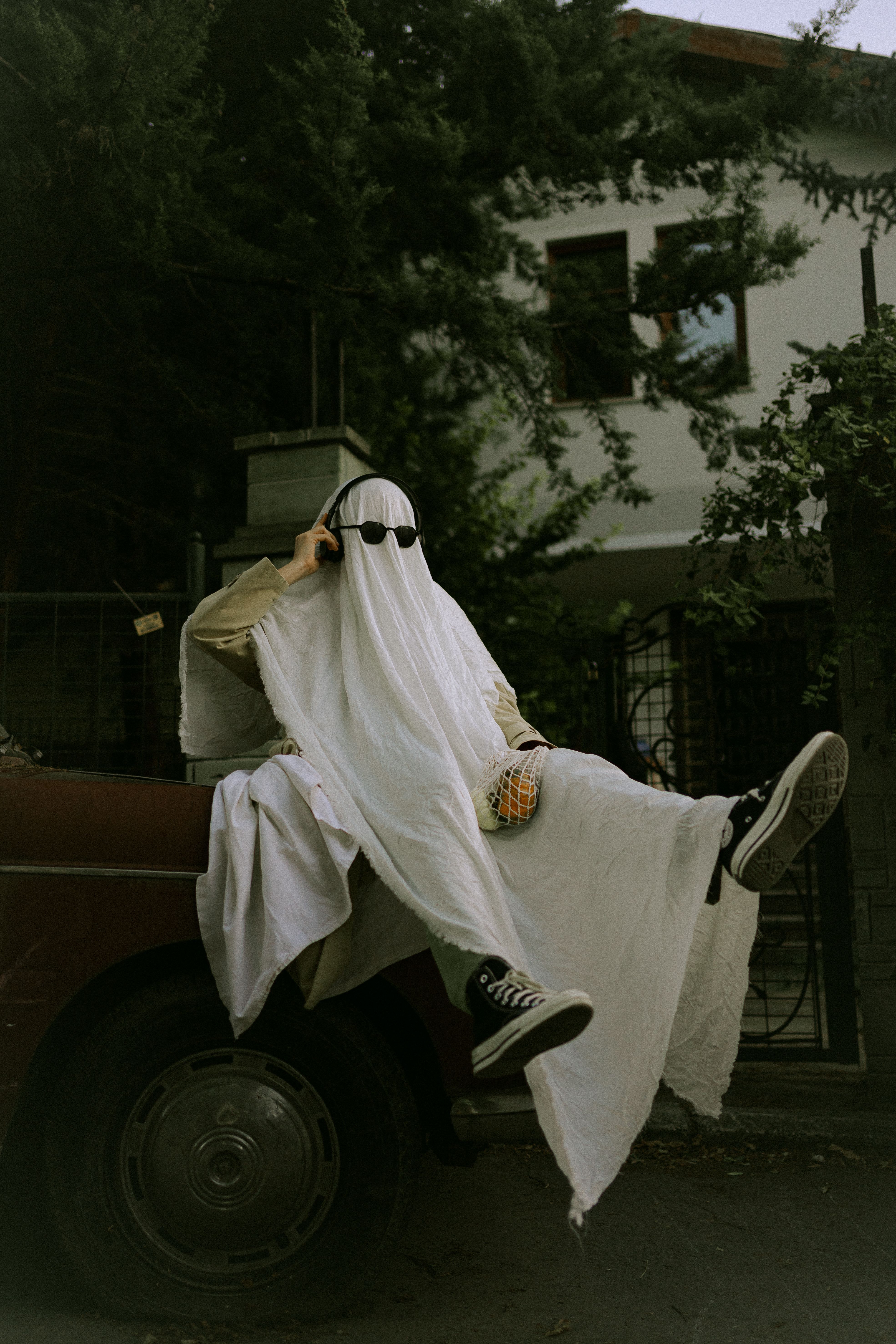 A person in a ghost costume with sunglasses sitting on a car, capturing a whimsical Halloween vibe.