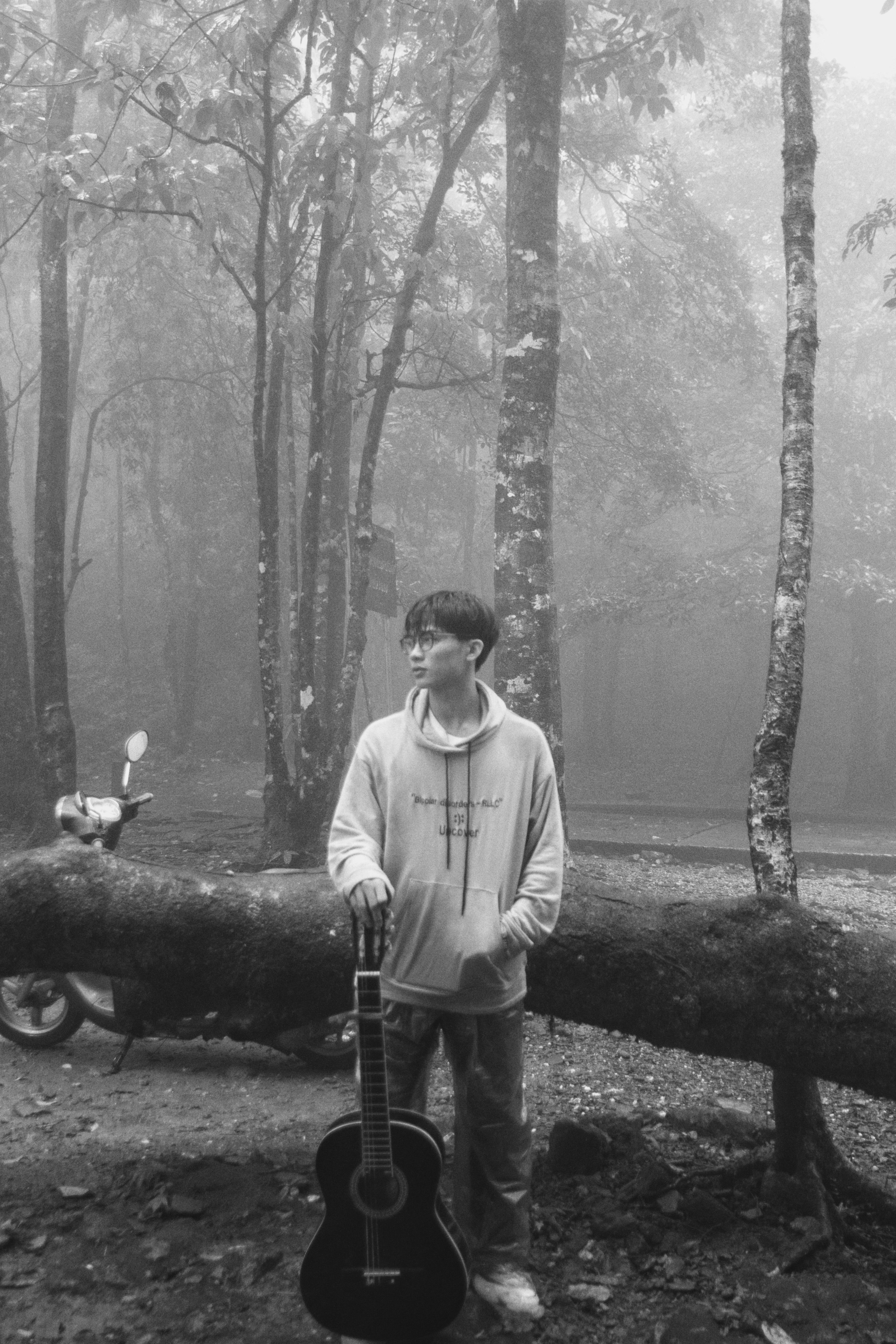 A young Asian man stands with a guitar in a misty forest environment.