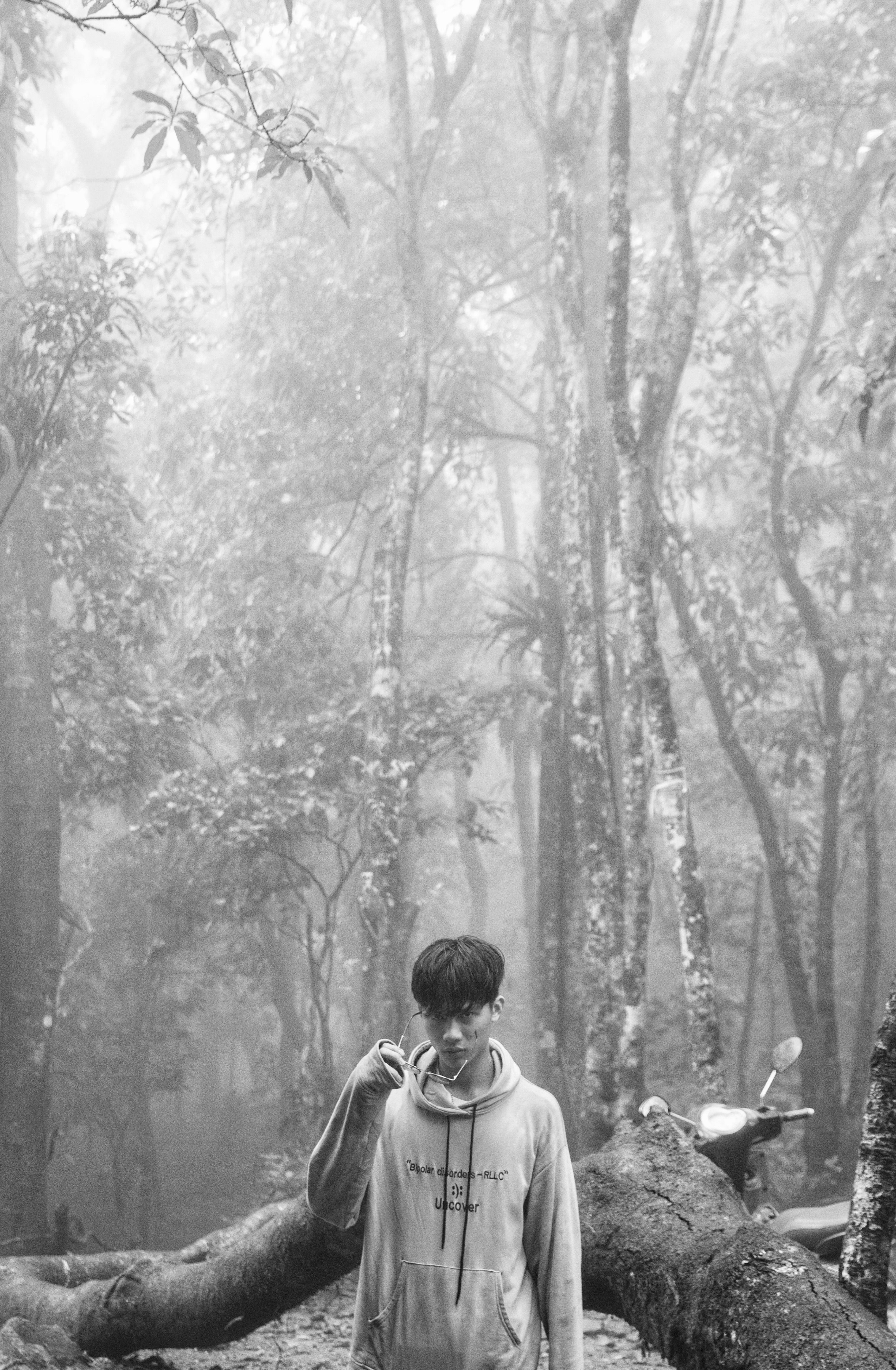 Black and white photo of a young man in a hoodie standing among misty forest trees.