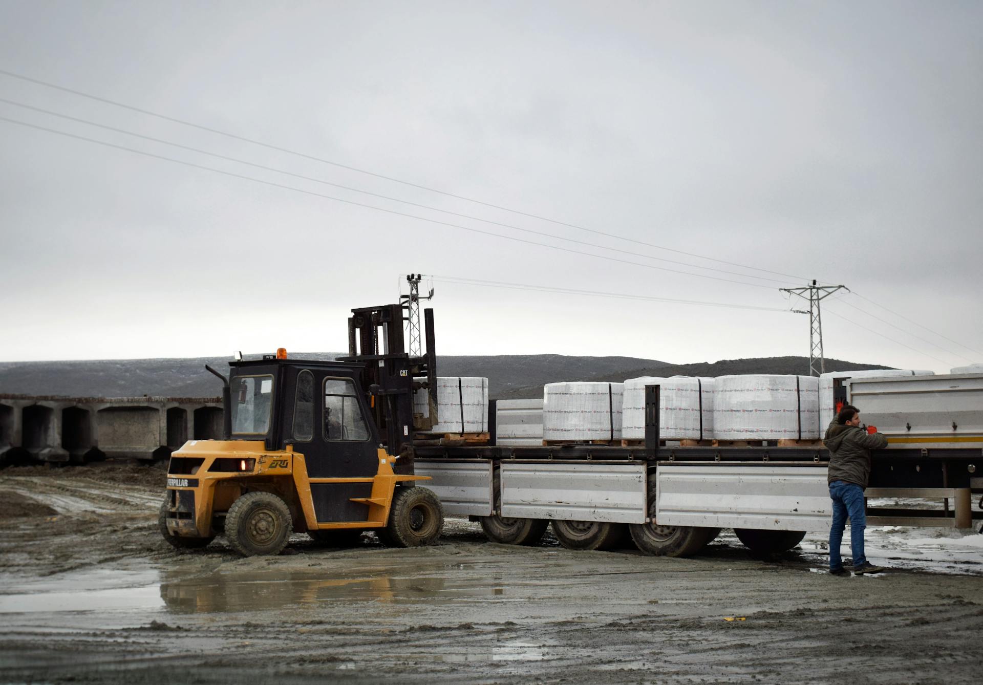 A forklift is loading cargo onto a truck in a muddy logistics area with a worker assisting.