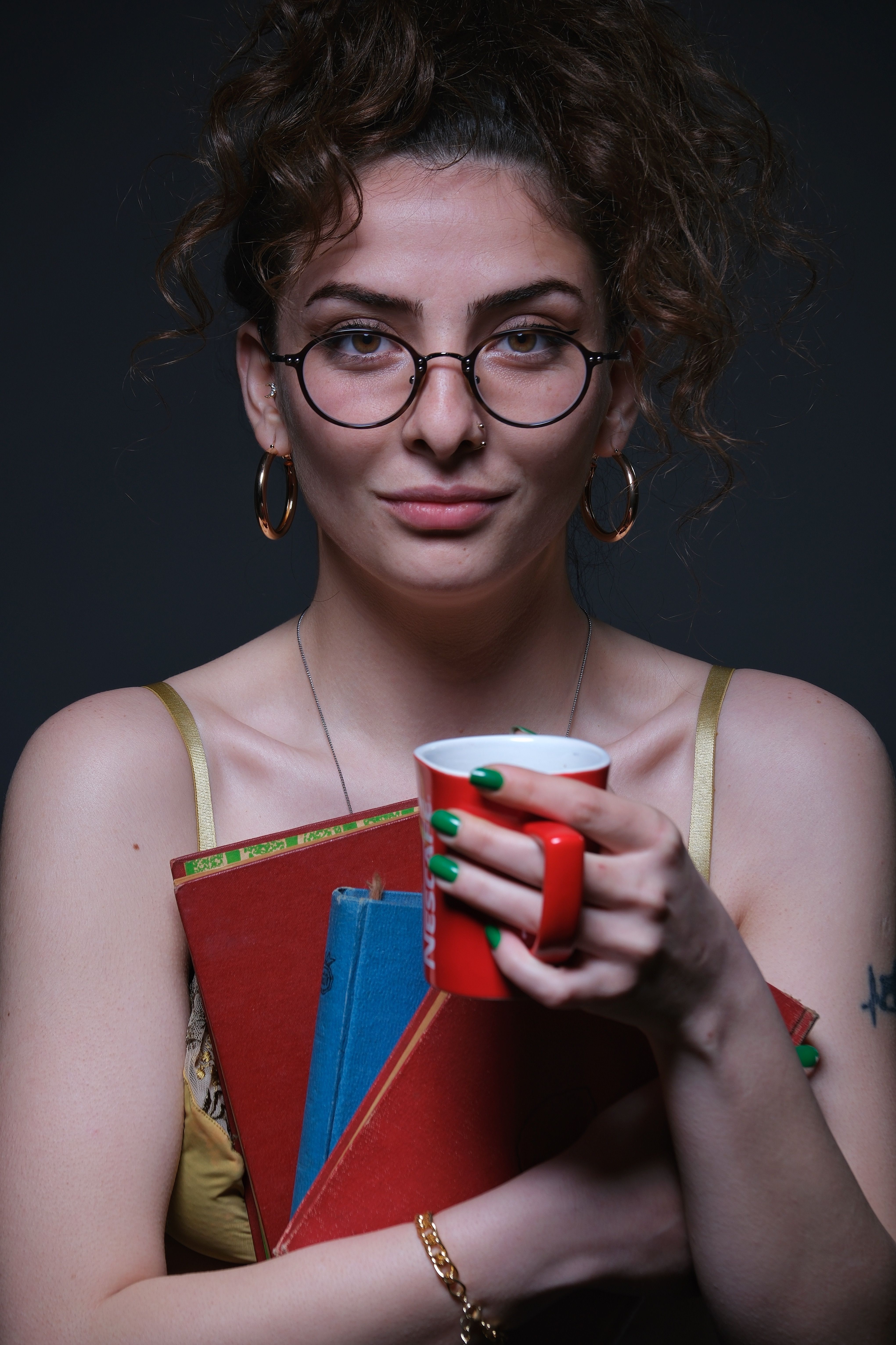 Curly-haired woman with eyeglasses holds books and coffee cup, smiling softly.