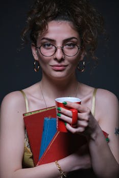 Curly-haired woman with eyeglasses holds books and coffee cup, smiling softly.