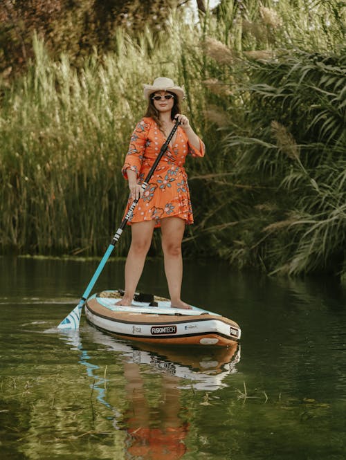 Woman in a Dress, Hat and Sunglasses Paddleboarding on a Body of Water 