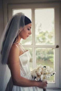 Beautiful bride in wedding dress holding a bouquet by a window
