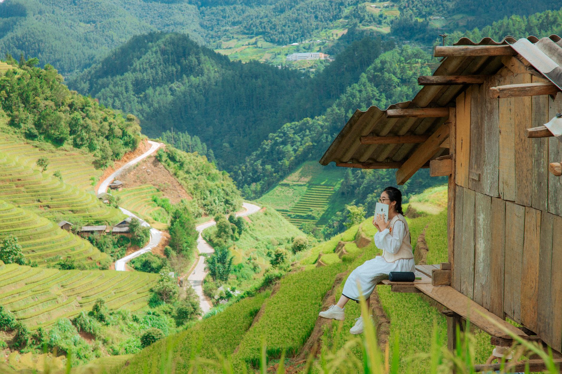 Asian woman reading a book while sitting on a hut, overlooking a lush green valley and terraced rice fields.