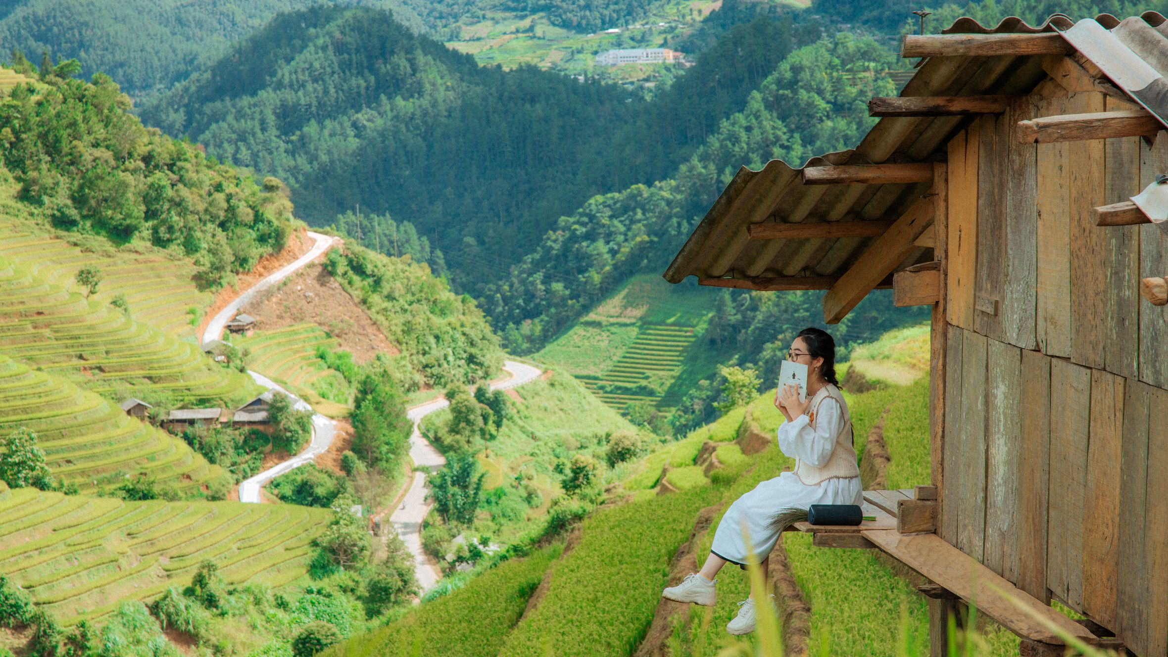 Asian woman reading a book while sitting on a hut, overlooking a lush green valley and terraced rice fields.