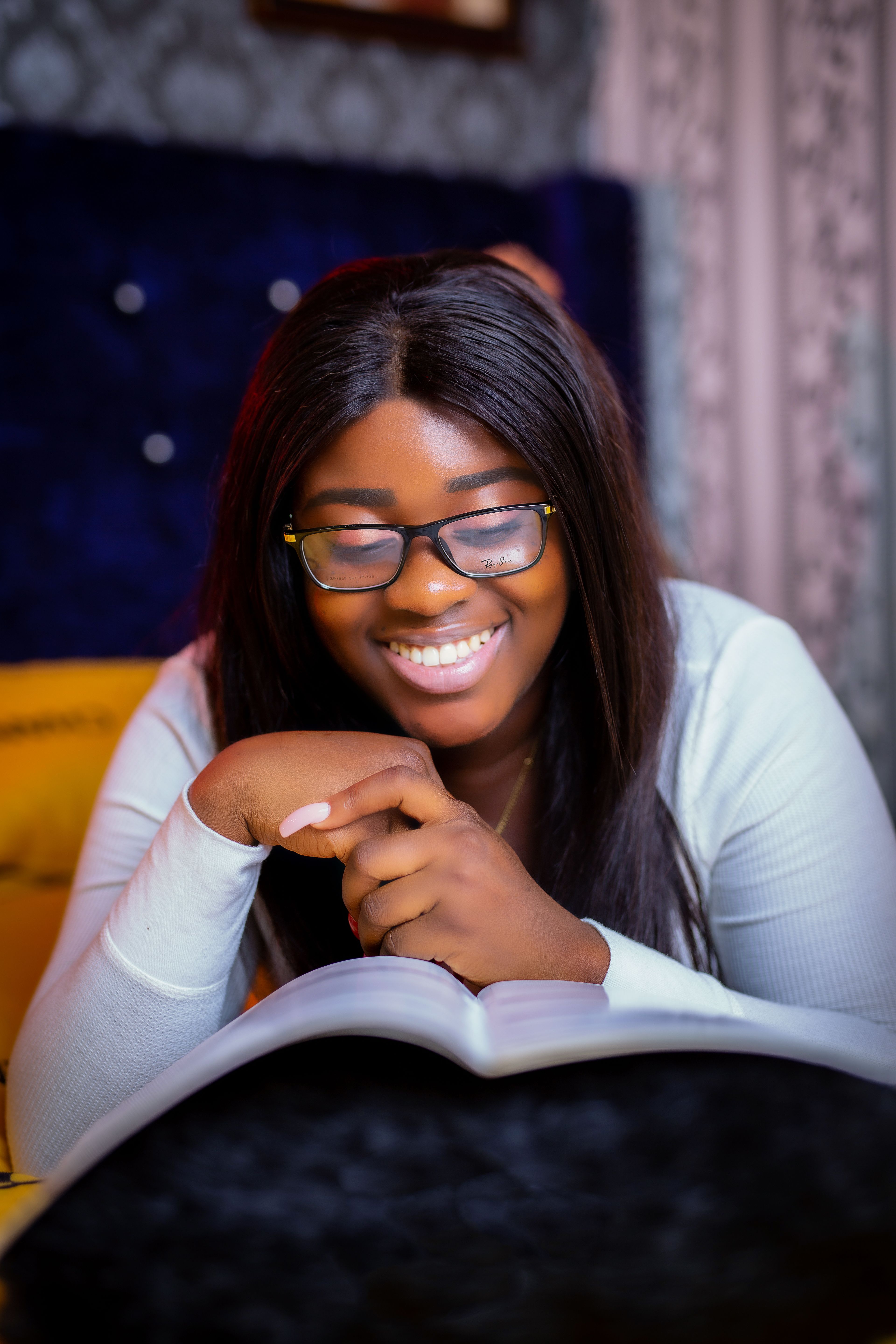 Smiling person with glasses reading an open book indoors, resting their chin on their hand