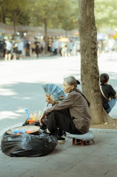 An elderly woman with gray hair sits selling items on a city sidewalk under the sunlight.