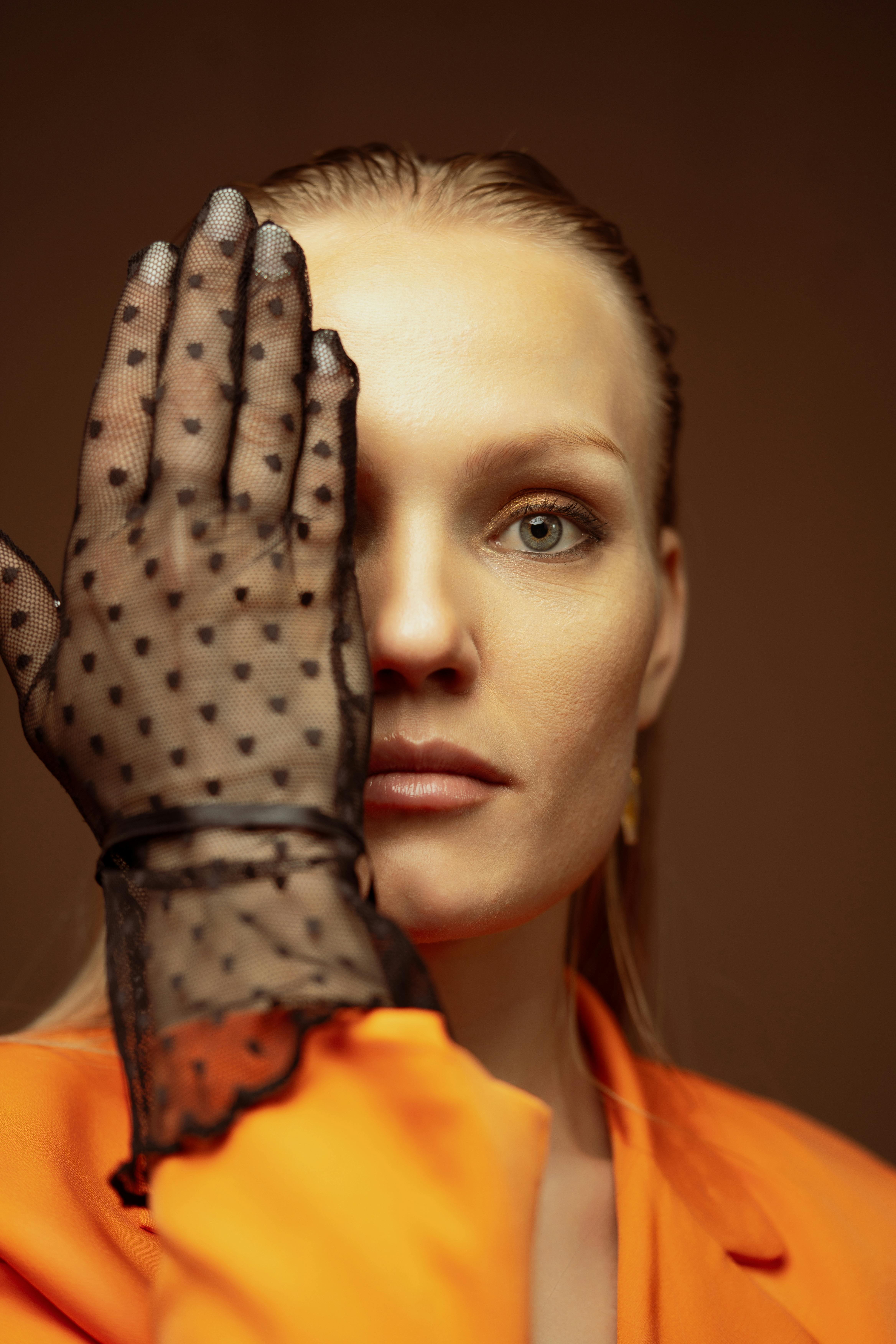 Stylish woman in orange with lace glove covering half her face, creating intrigue.