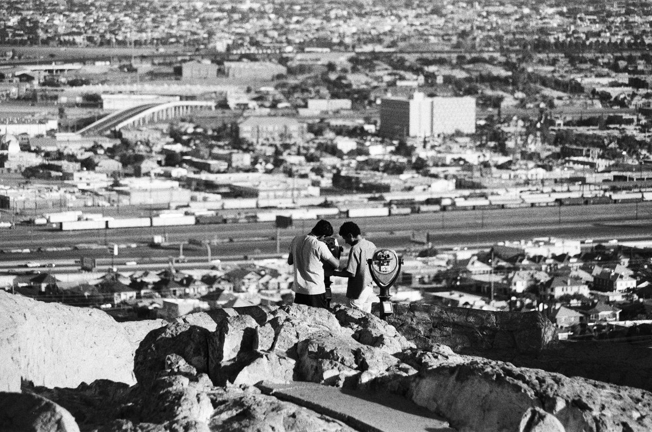 black and white photo of two men observing a city from a rocky hill vi