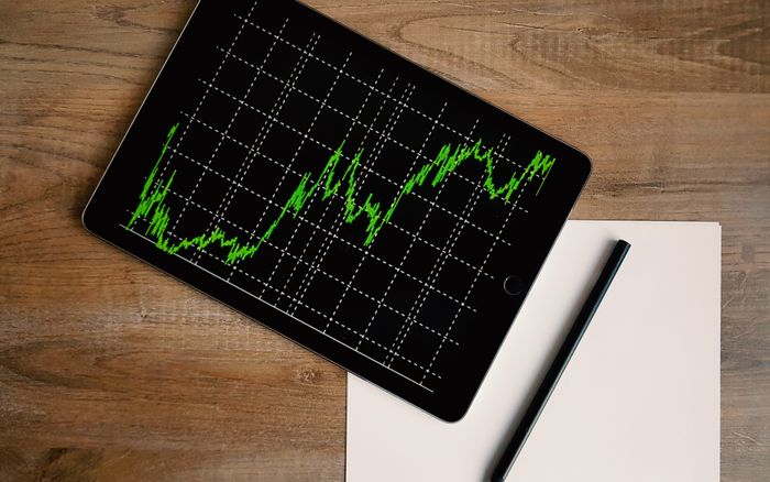 An iPad displaying a stock market graph on a wooden desk beside a pencil and paper.