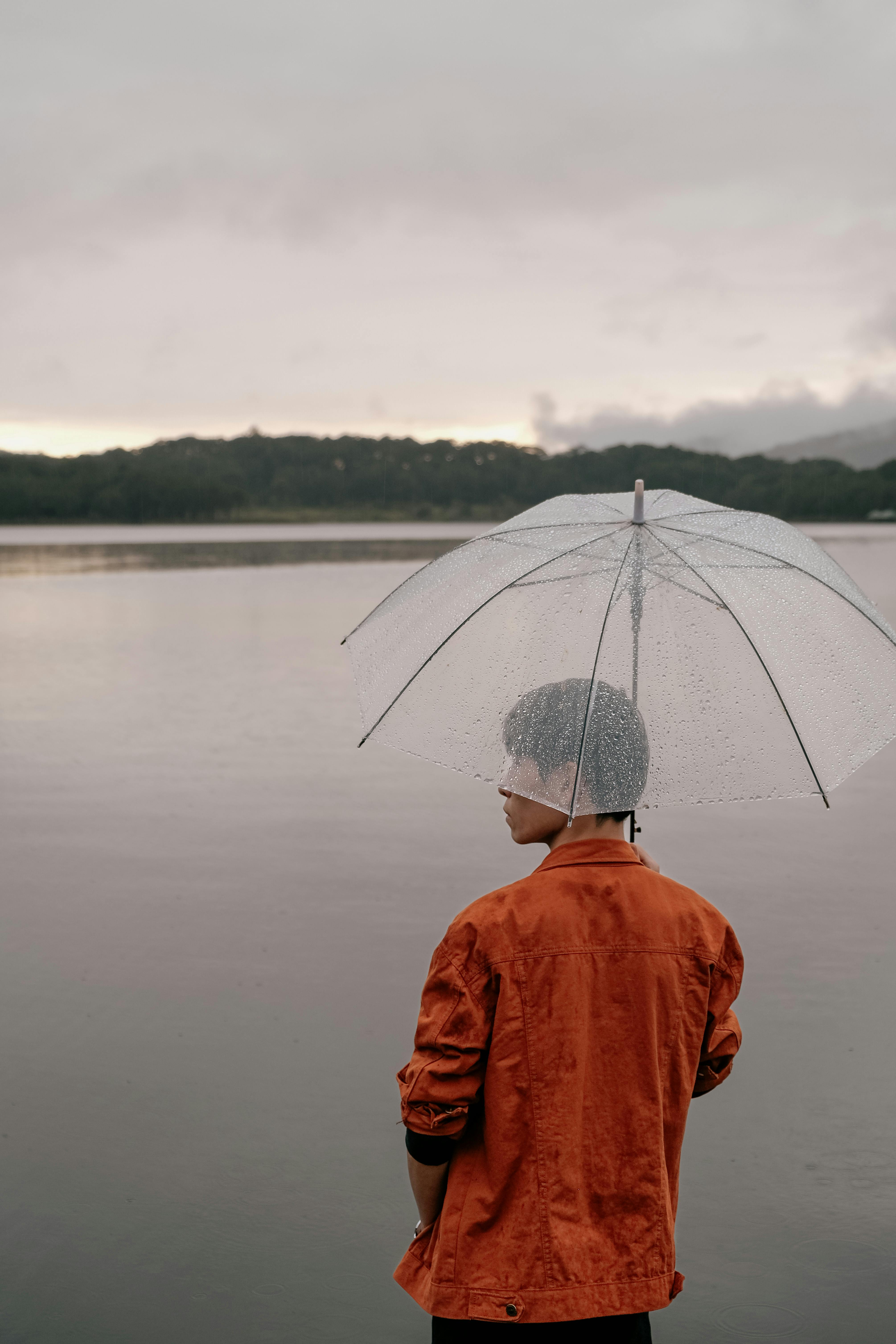 A man in an orange jacket stands by a lake in Dalat, Vietnam, holding a clear umbrella as rain falls gently.
