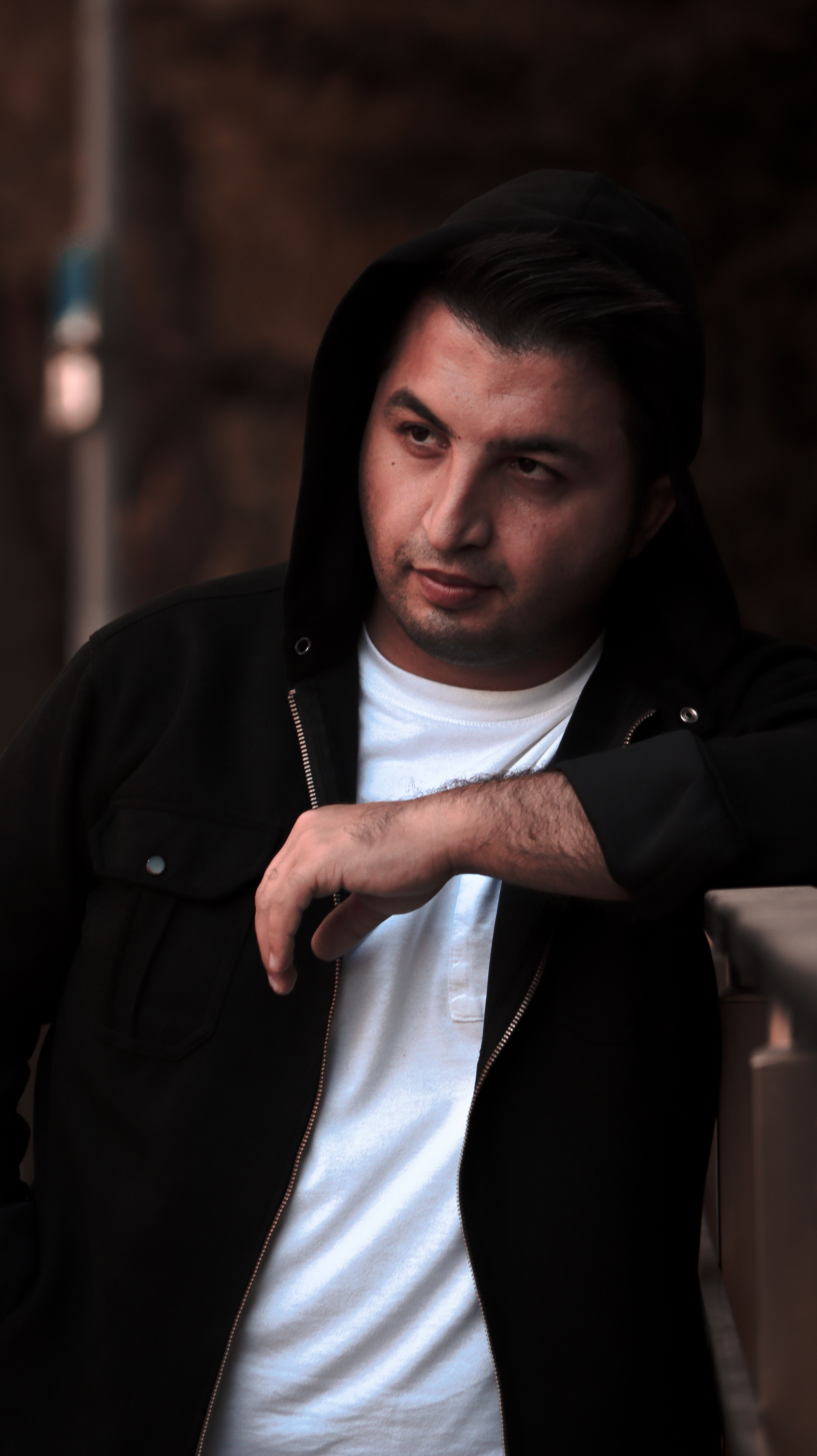 Portrait of a man in black hoodie leaning on a railing outdoors, exuding a moody vibe.