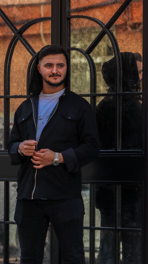 Free A young man with a beard and mustache poses confidently in a black jacket against a reflective surface. Stock Photo