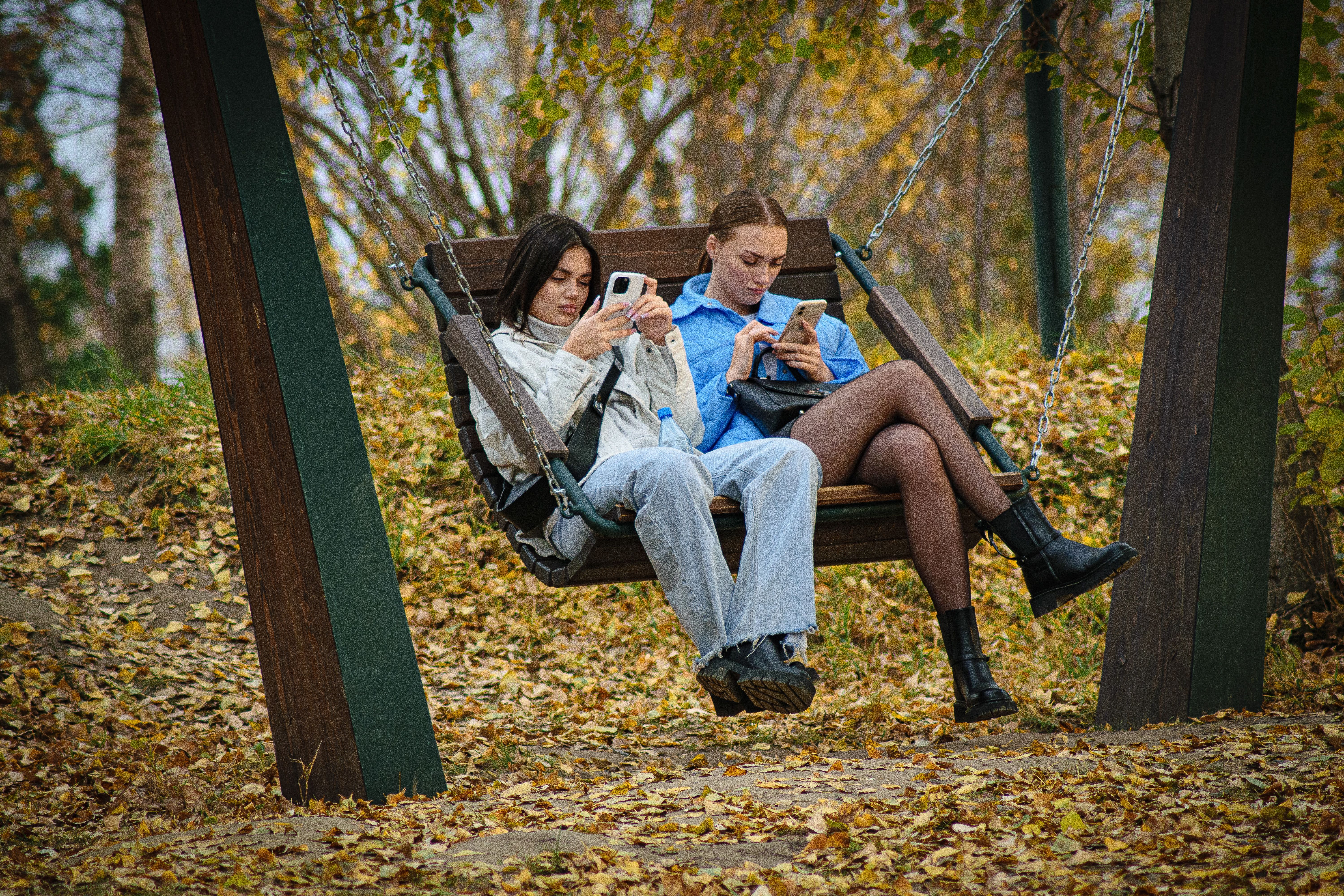 Two young women sitting on a swing using smartphones, surrounded by autumn leaves in a park.