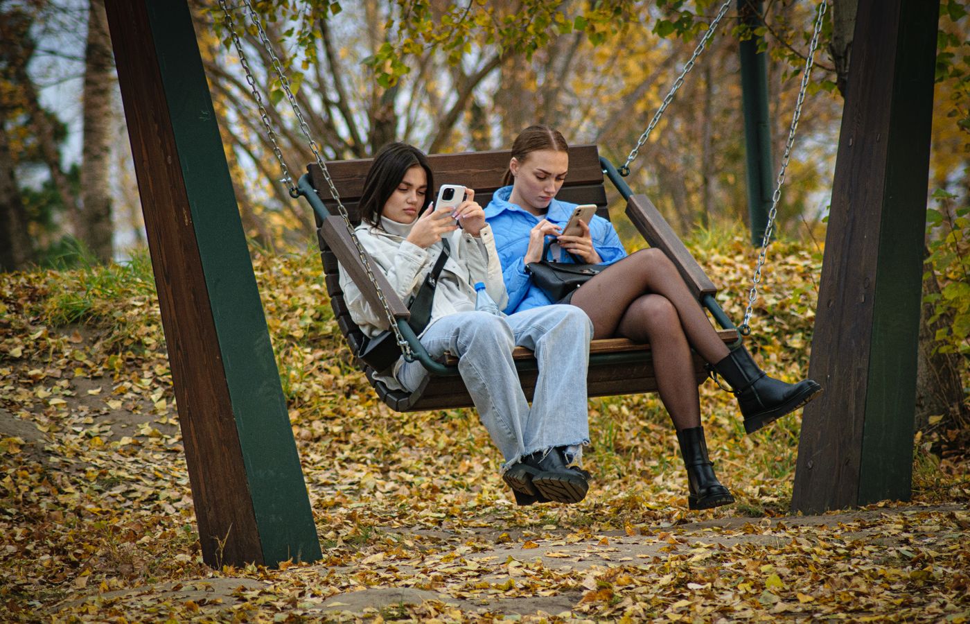 Two young women sitting on a swing using smartphones, surrounded by autumn leaves in a park.