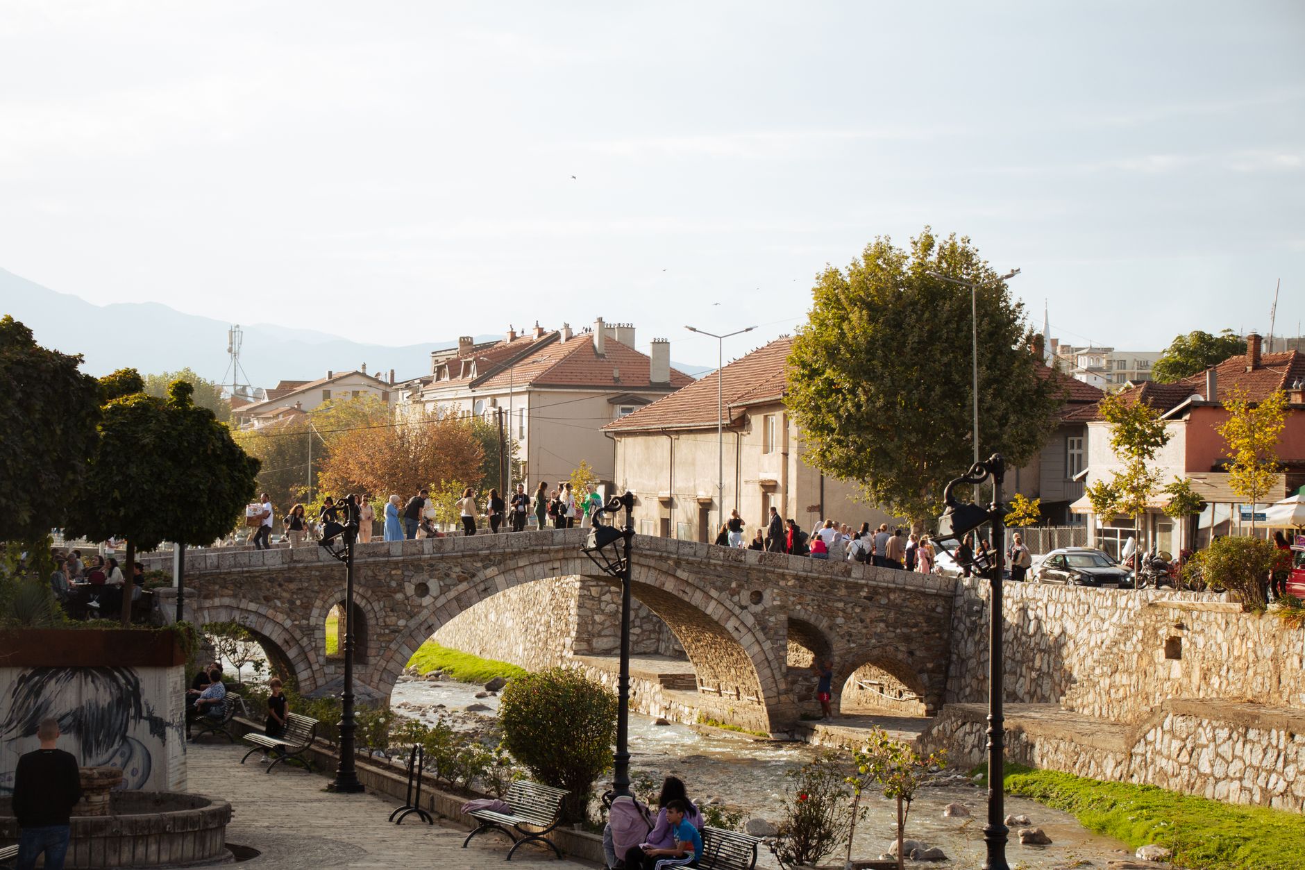 Scenic view of a historic stone bridge over a river in Prizren, Kosovo, with people walking and a charming urban backdrop.. Photo by Sabina Kallari on Pexels