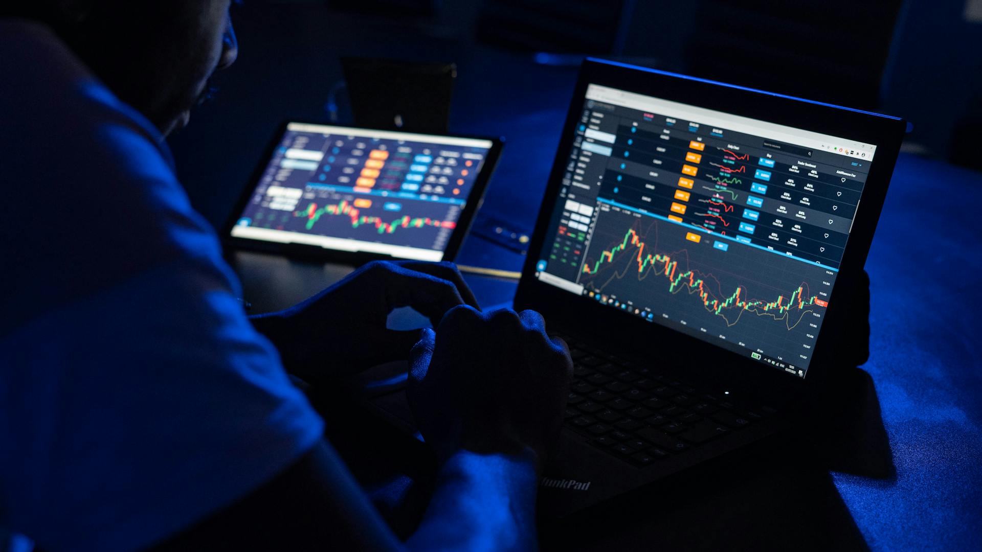 Man analyzing financial charts and data on laptops in a dimly lit room, highlighting forex trading.