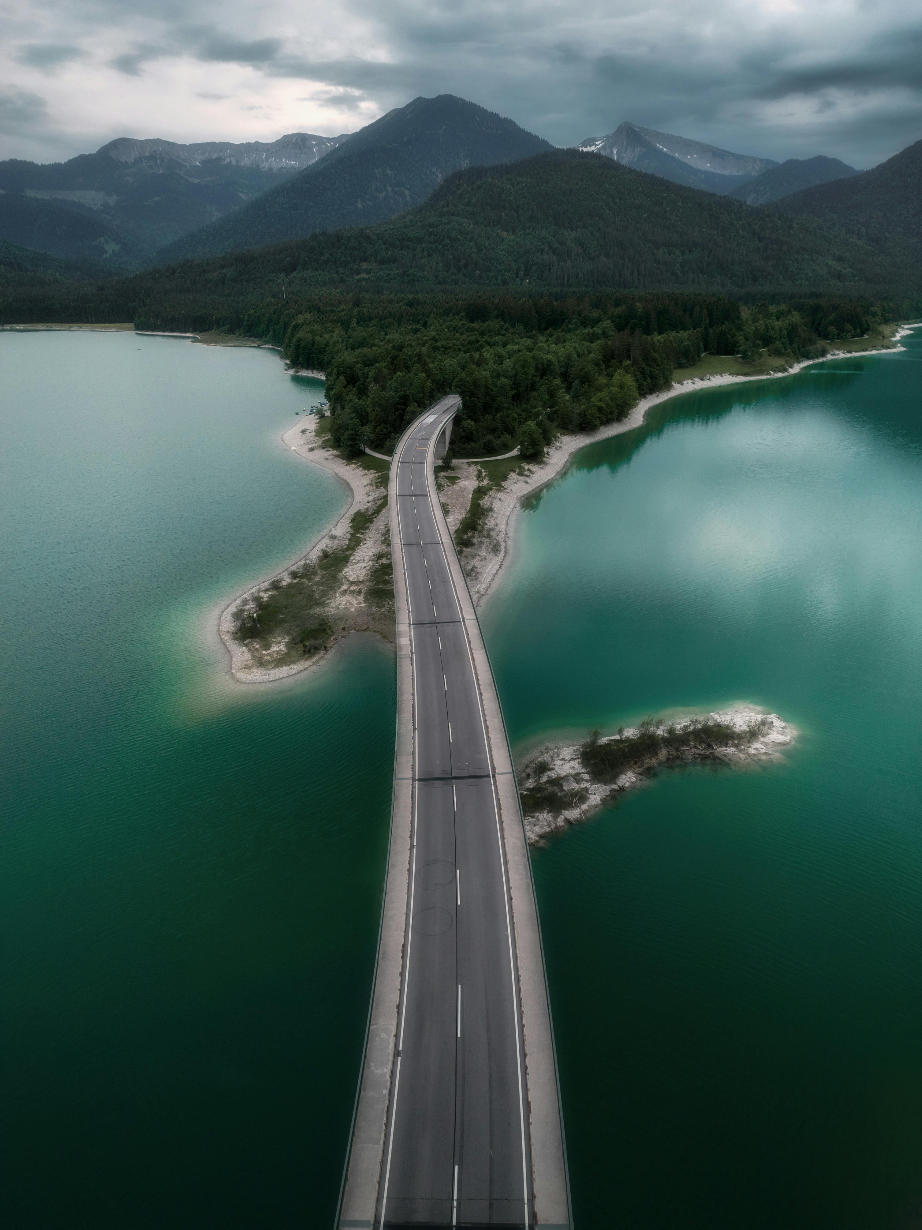 Aerial view of a bridge crossing between vibrant teal lakes and lush green forests with mountains in the background.