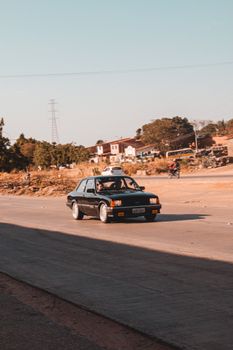A retro black sedan driving on an isolated road in a rustic town during sunset.