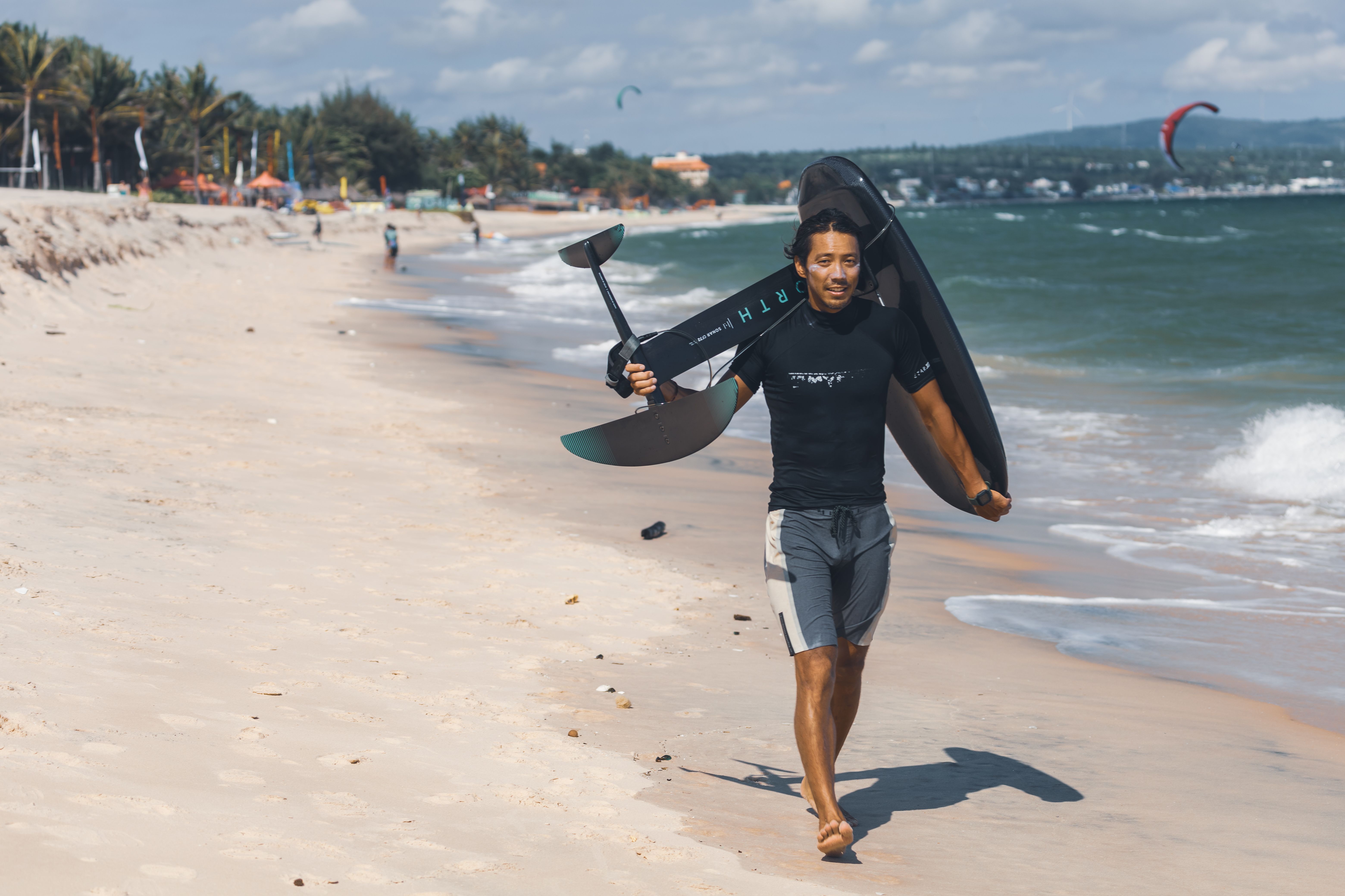 A kitesurfer carrying a hydrofoil board on a sunny beach, highlighting summer beach sports.