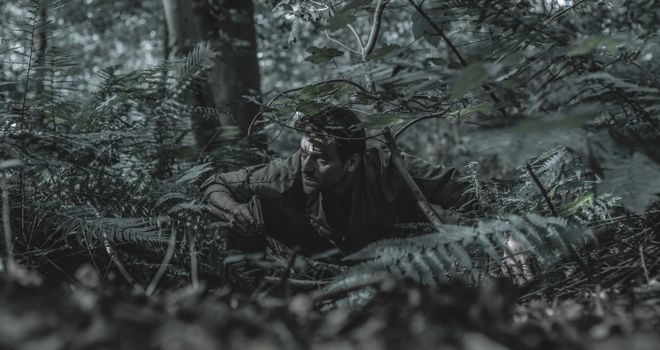 A man crouches in dense forest foliage, blending with his surroundings, evoking a sense of adventure and stealth.