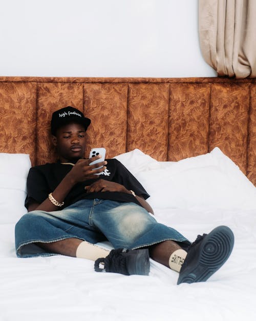 Man Wearing T-shirt Loose Jeans and Trainers Texting Lying on Bed in Hotel Room