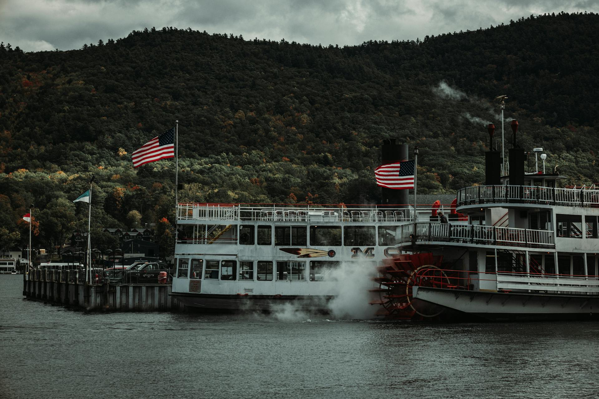 Steamboat with American flags docked on a serene lake with autumn-colored mountains in the background.