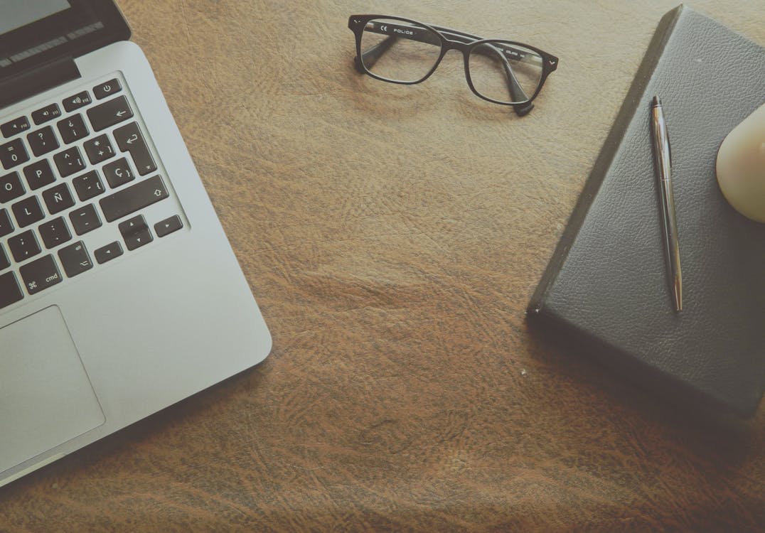 Close up of a desk featuring a laptop, pair of glasses, pen and a black leather journal.