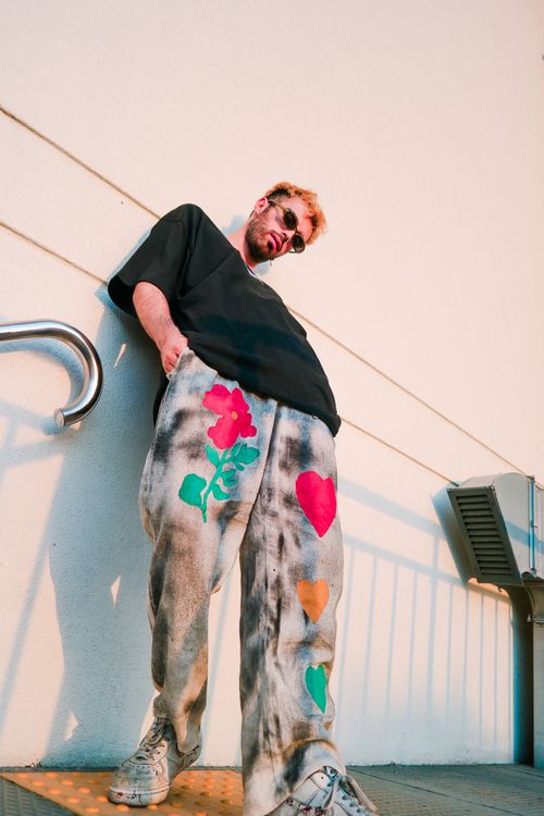 Free A fashionable man wearing artistic pants with bold patterns, leaning against a wall outdoors. Stock Photo