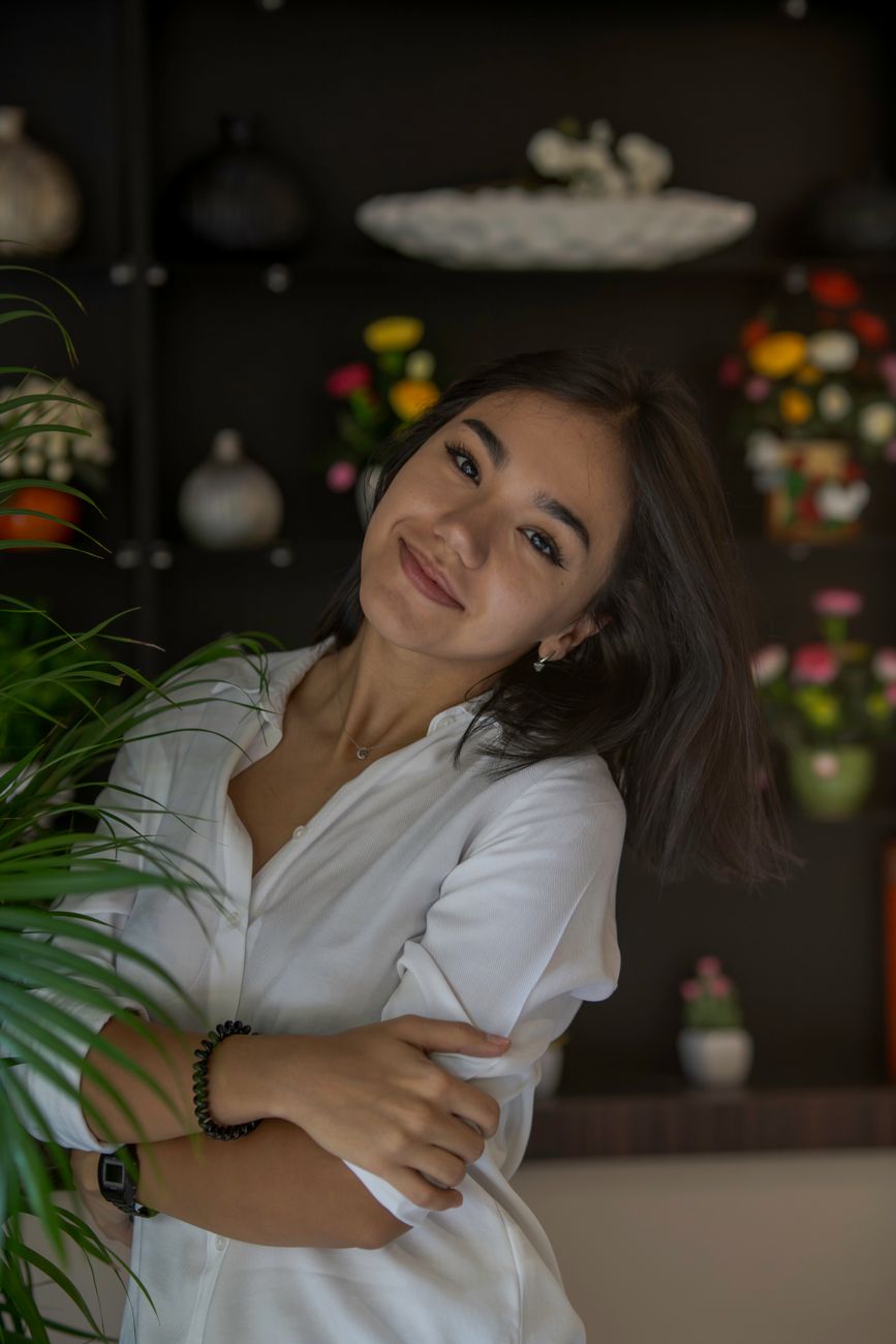 Cheerful woman in white shirt smiling and posing indoors with vibrant plant decor.