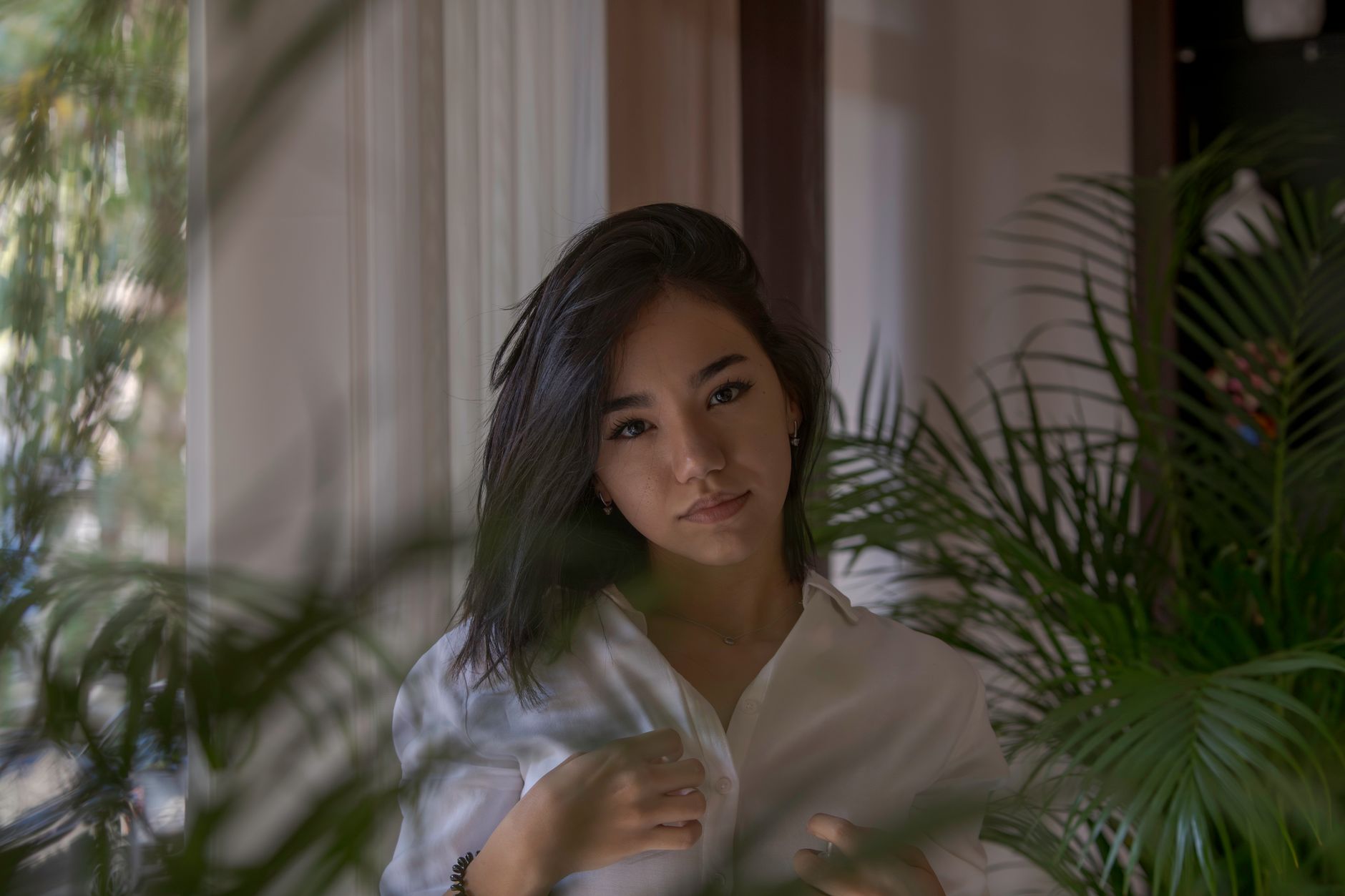 A young woman wearing a white blouse stands indoors among lush green plants, casting a serene mood.
