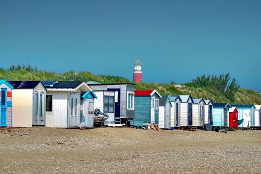 A row of vibrant beach huts along a sandy shore with a lighthouse in the distance under a clear blue sky.