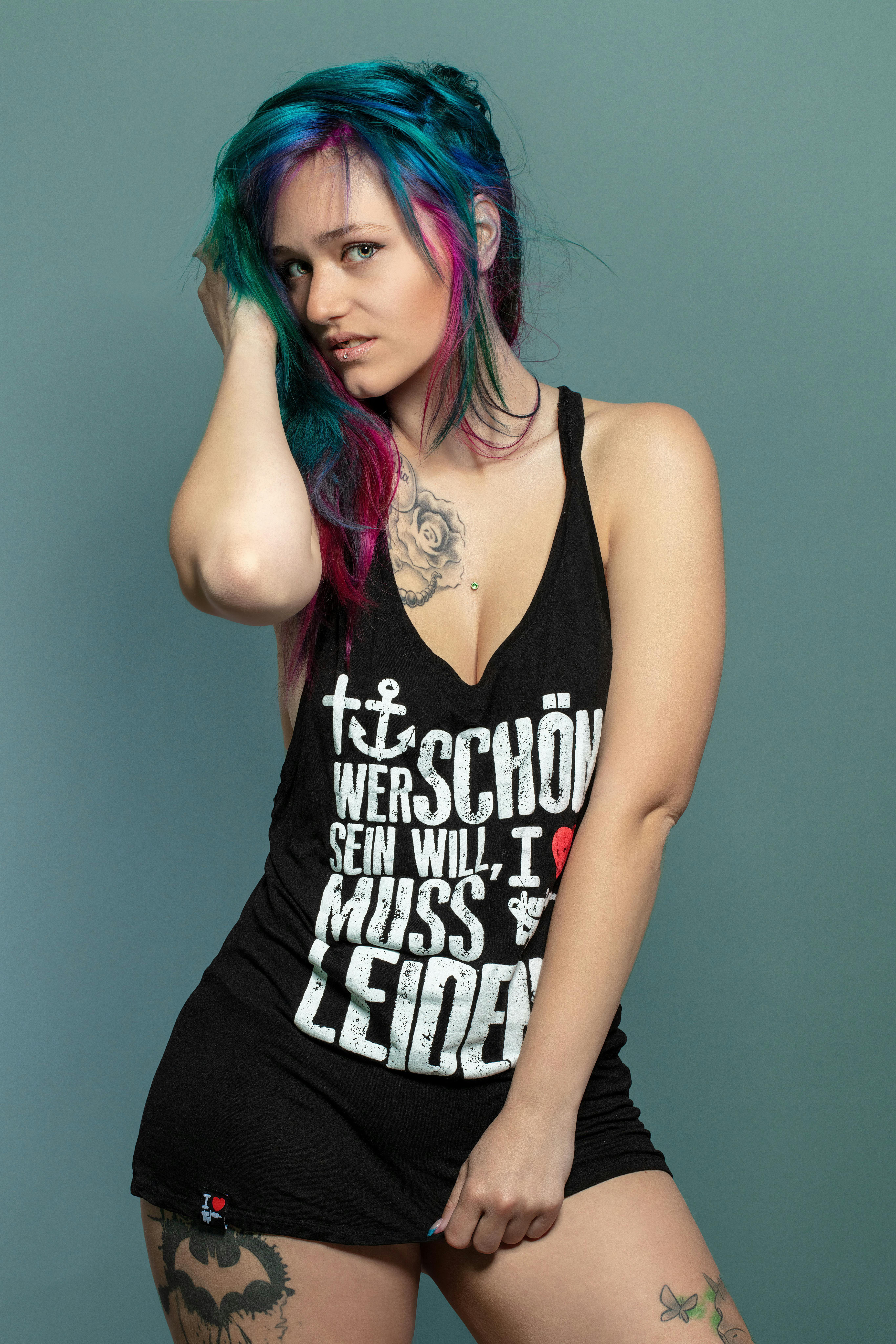 Woman in black tank top with tattoos, colorful hair, and expressive pose indoors.