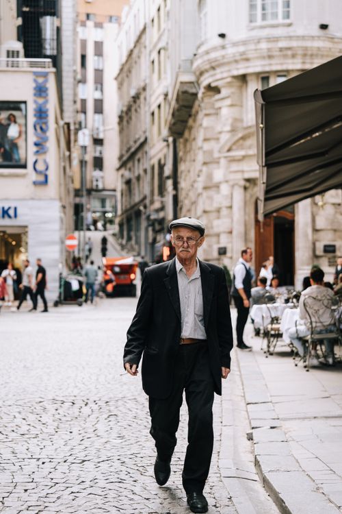 Free An elderly man with eyeglasses and a mustache walks down a cobblestone city street past urban cafes. Stock Photo
