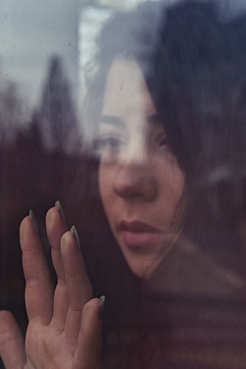 Free A thoughtful young woman looks out a window, her reflection showing emotion. Stock Photo