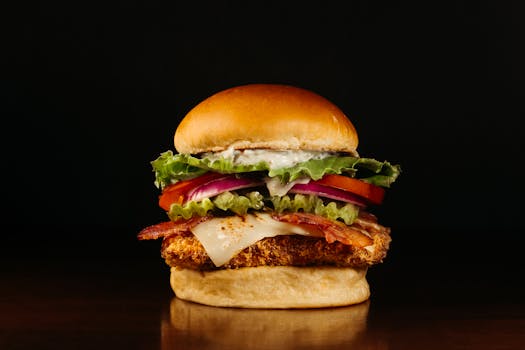 Tasty chicken burger with bacon, cheese, and fresh veggies in a bun against a dark backdrop.