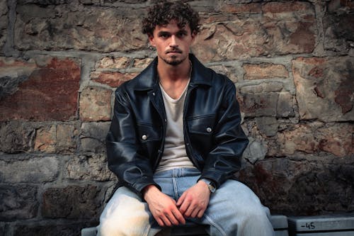 Male Model Wearing a Black Leather Jacket Sitting by a Stone Wall