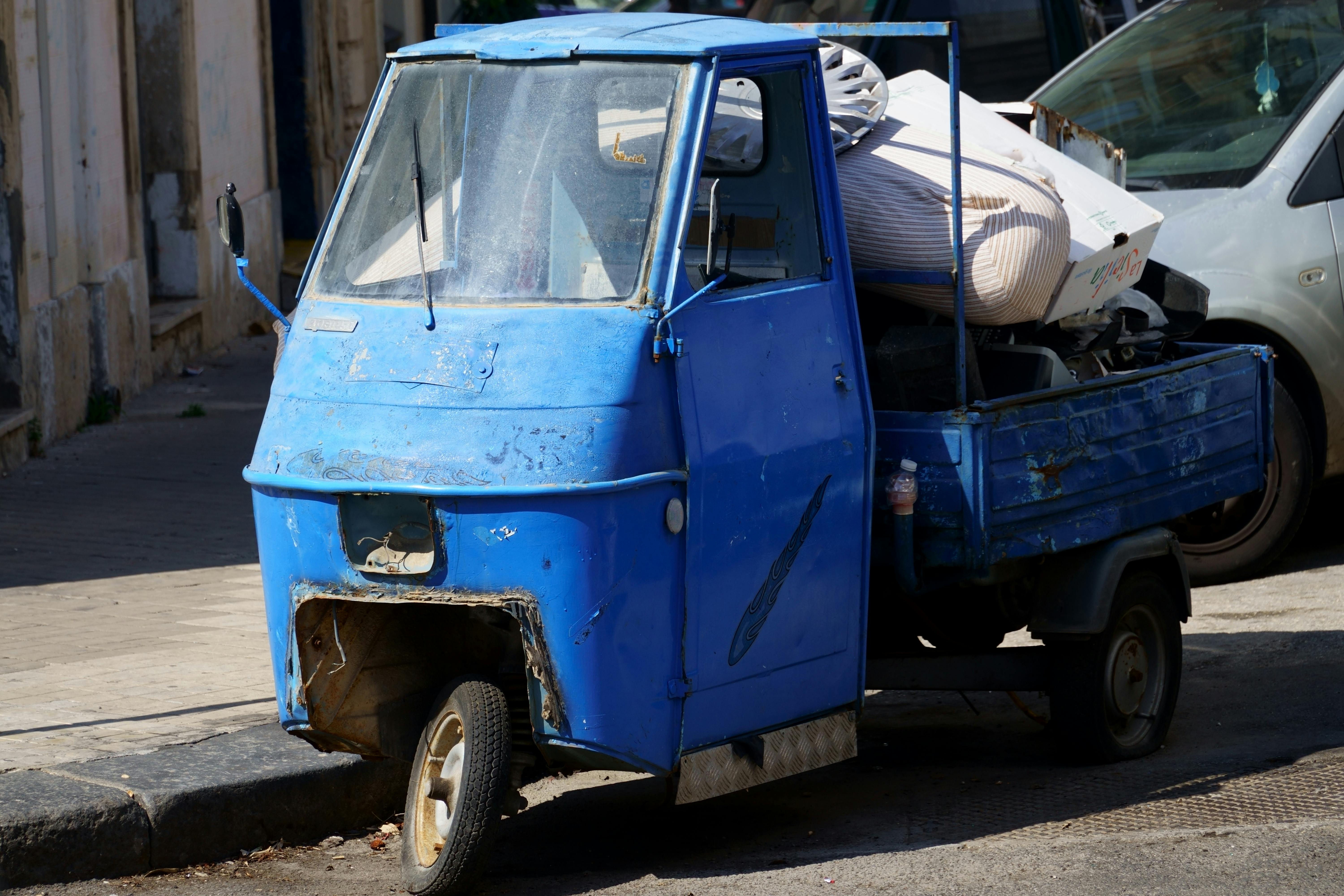 Free A weathered blue three-wheeler rickshaw parked on a city street, filled with clutter. Stock Photo